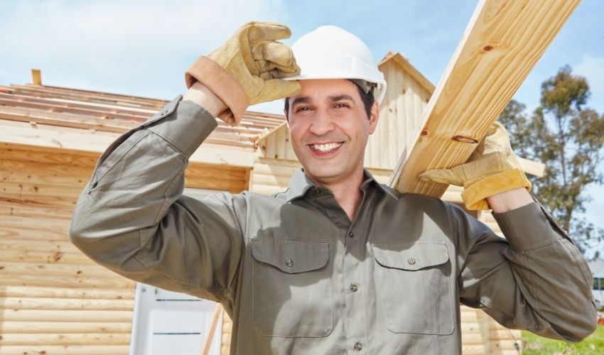 A smiling construction worker in a hard hat and gloves carries a wooden plank on his shoulder, standing in front of a wooden house under construction—ideal for those exploring kanada-experten-migration on a sunny day.